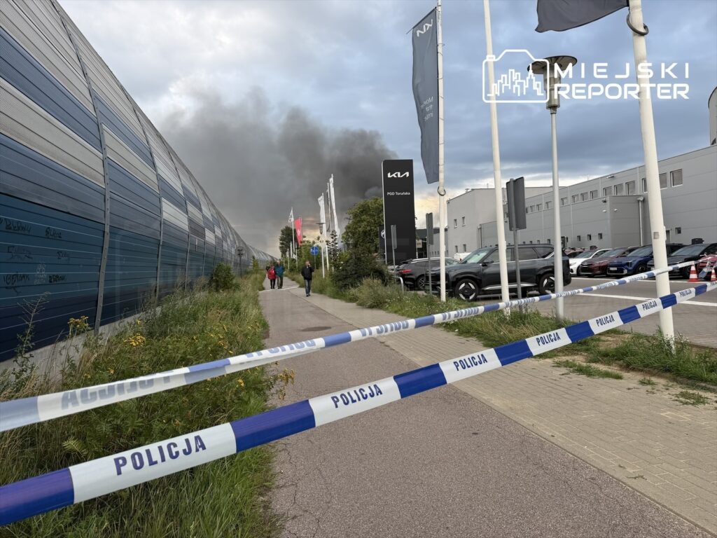 Słupki policyjne oddzielają przechodniów od strefy pożaru, w tle widoczne kłęby dymu wydobywające się z hali.
