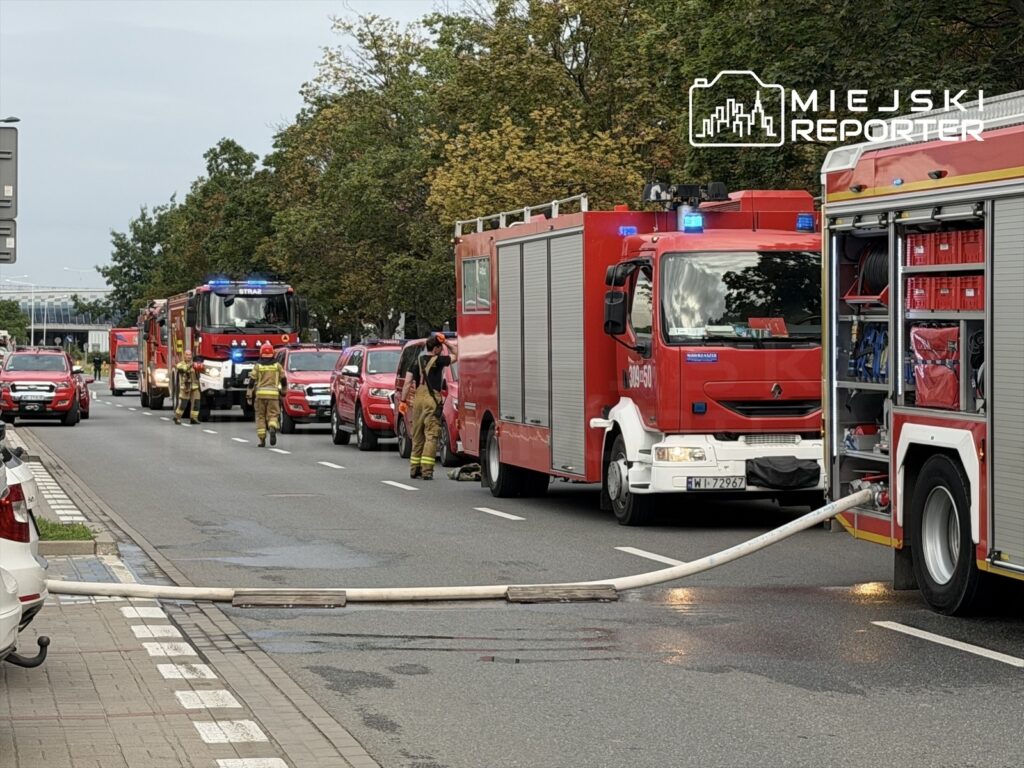 Strażacy w mundurach pracują przy wozach strażackich na ulicy, prowadząc działania gaśnicze z użyciem węża.