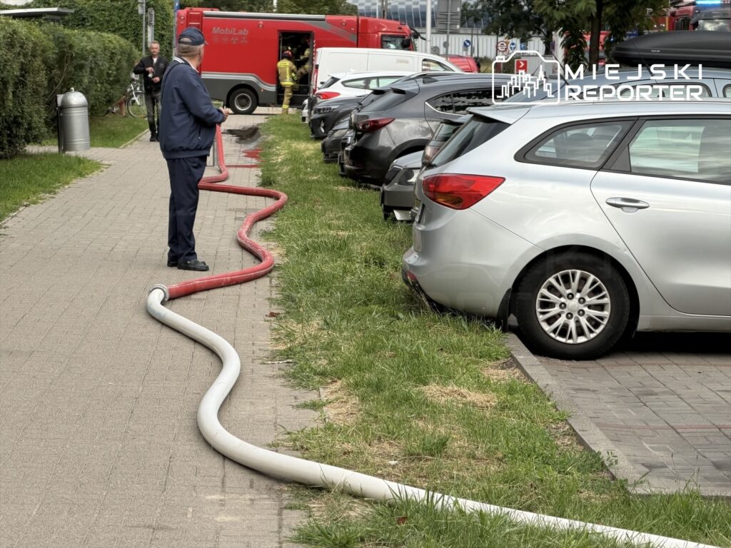 Mężczyzna w mundurze trzyma wąż strażacki na chodniku obok zaparkowanych samochodów, w tle widać pojazdy ratunkowe.