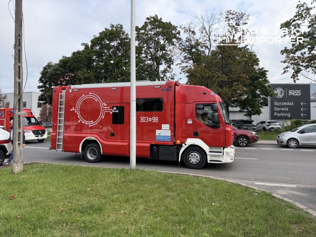 Czerwony wóz strażacki z numerem 303 w98B przejeżdża obok parkingu i serwisu samochodowego w mieście.