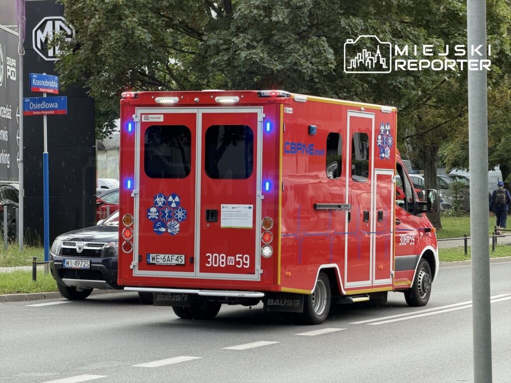Czerwony ambulans z włączonymi sygnałami świetlnymi przejeżdża obok znaku ulicy Krasnobrodzkiej.