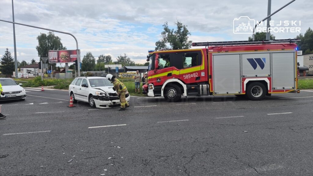 Strażak sprawdzający uszkodzenia białego samochodu osobowego na drodze, obok wozu strażackiego.