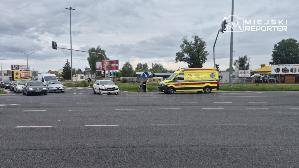 Na skrzyżowaniu stoi uszkodzony samochód osobowy, obok niego na poboczu znajduje się karetka pogotowia oraz kilka osób.