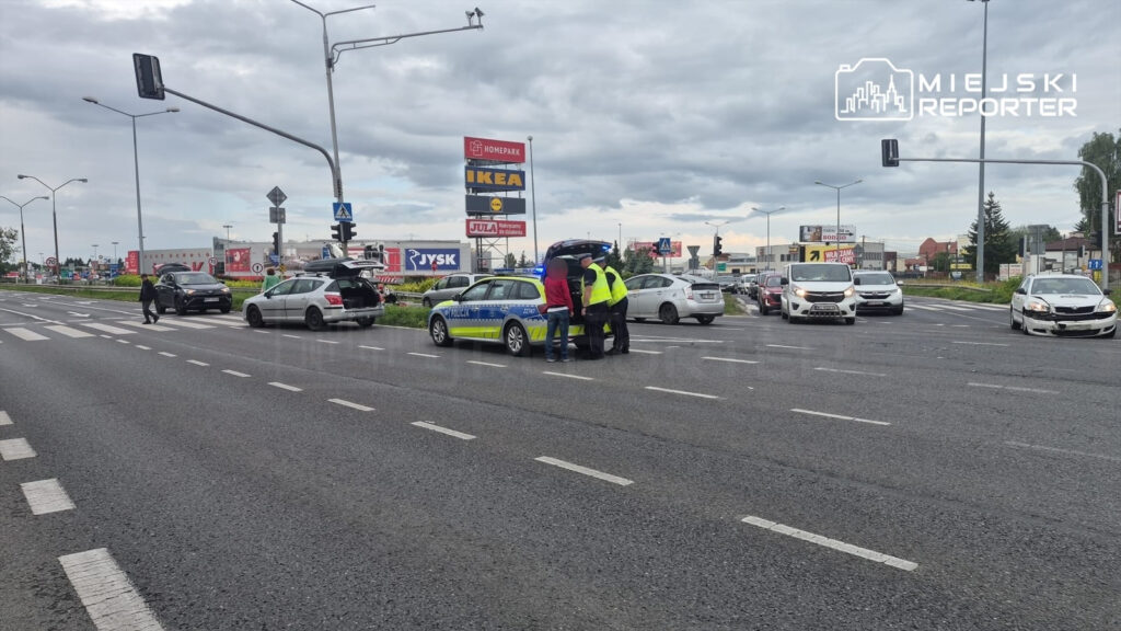 Policjanci rozmawiają przy radiowozie na ruchliwej drodze, obok zatrzymane samochody osobowe. W tle widoczne reklamy.