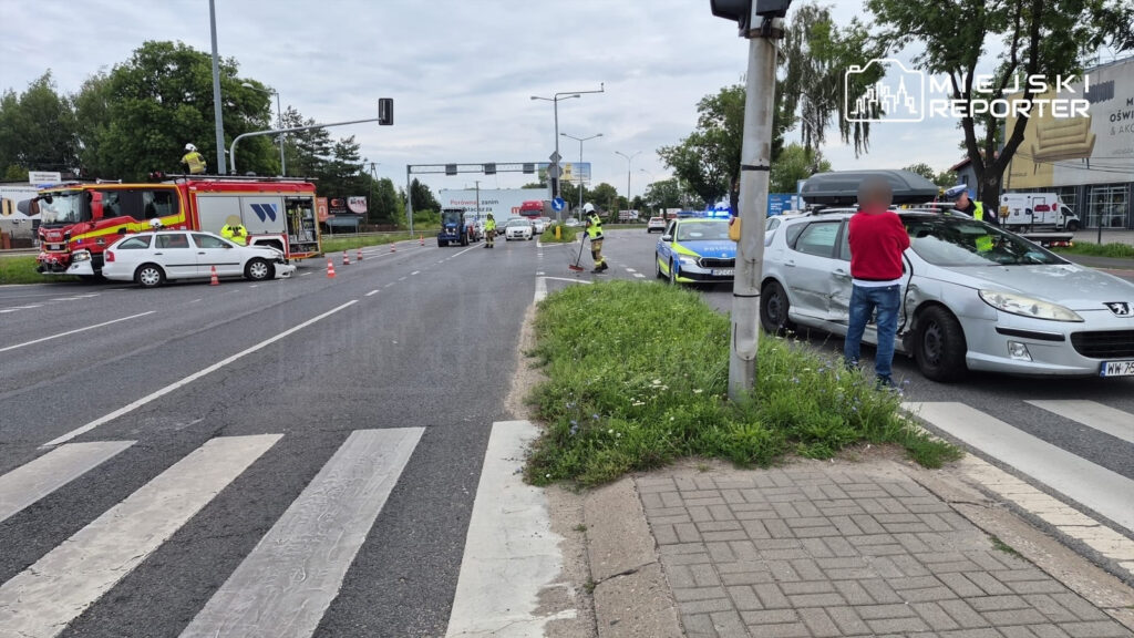 Na jezdni stoi uszkodzony samochód osobowy, obok niego pracują strażacy i policjanci; w tle widoczne pojazdy ratunkowe.