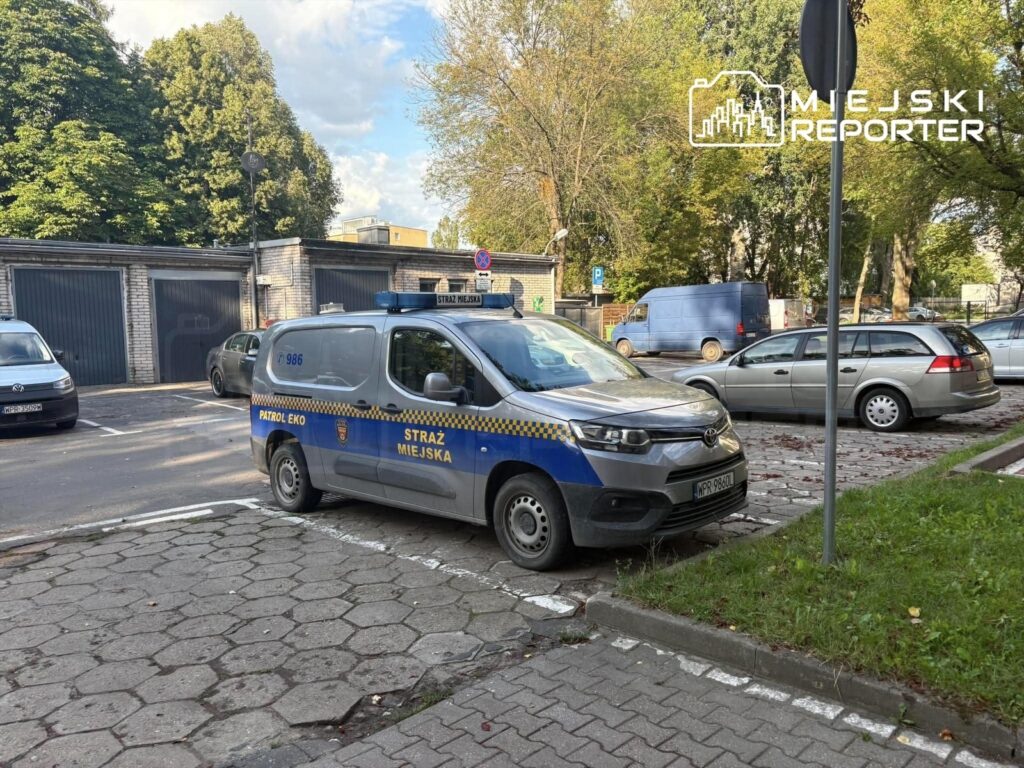Samochód straży miejskiej zaparkowany na chodniku w pobliżu garaży, otoczony innymi pojazdami.