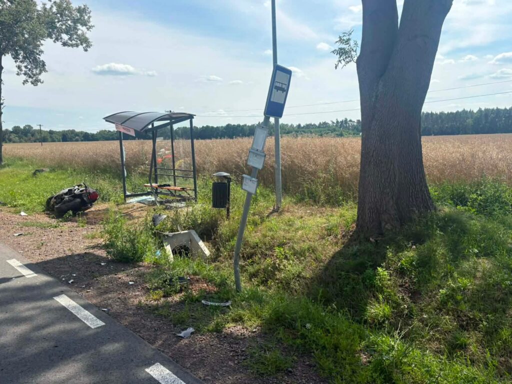 Przystanek autobusowy z wiatą w polu, obok leży przewrócony motor, w tle widoczne zboże i drzewa.