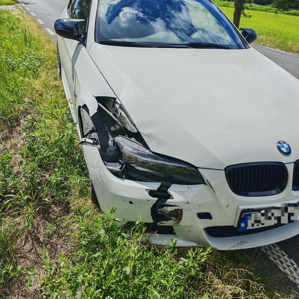 Uszkodzony biały samochód marki BMW zaparkowany na poboczu drogi, z wgniecionym przodem i uszkodzoną lampą.