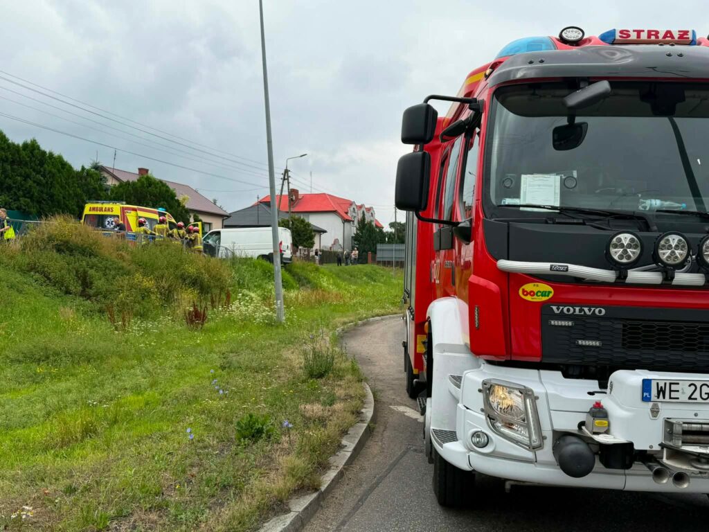 Czerwony wóz strażacki zatrzymany przy drodze, obok zespołu ratowników medycznych i ambulansu w tle.