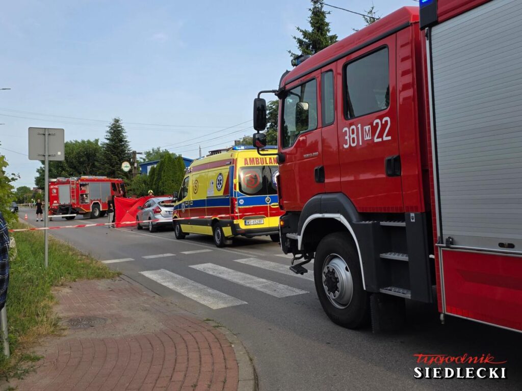 Na drodze zablokowanej przez służby ratunkowe stoi czerwony wóz strażacki i ambulans, a w tle widoczne są policyjne taśmy.
