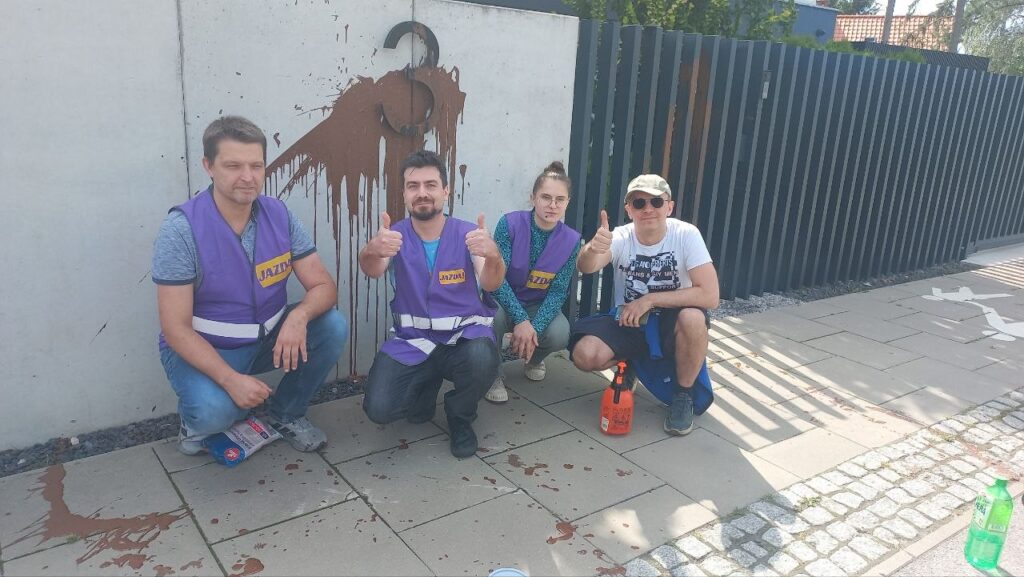 Czterech wolontariuszy w fioletowych kamizelkach uśmiecha się i pokazuje kciuki w górę przed zniszczoną ścianą z graffiti.