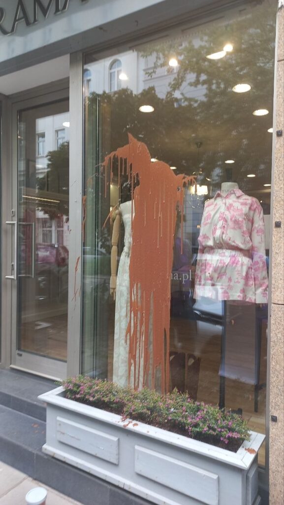Skrzynka z kwiatami przed witryną sklepu z odzieżą, na której spływa brązowa farba, zasłaniając manekiny w sukienkach.