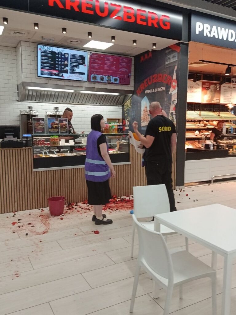 Kobieta w fioletowej kamizelce rozmawia z mężczyzną w czarnej koszulce w fast foodzie, na podłodze leżą resztki jedzenia.