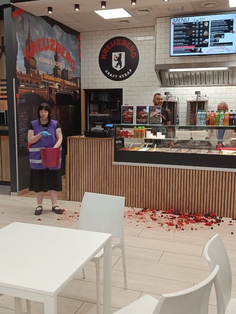Kobieta w fioletowej kamizelce trzymająca wiadro, sprząta strzępy jedzenia na podłodze w lokalu gastronomicznym.