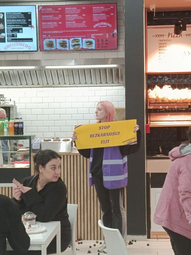 Kobieta z różowymi włosami trzyma transparent z napisem "STOP BEZKARNOŚCI ELIT" w restauracji fast food.