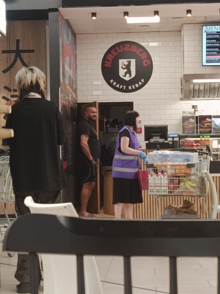 Kobieta w fioletowej kamizelce rozmawia z mężczyzną w koszulce, stojąc przy ladzie kebabowej. W tle widać zakupy w wózku.