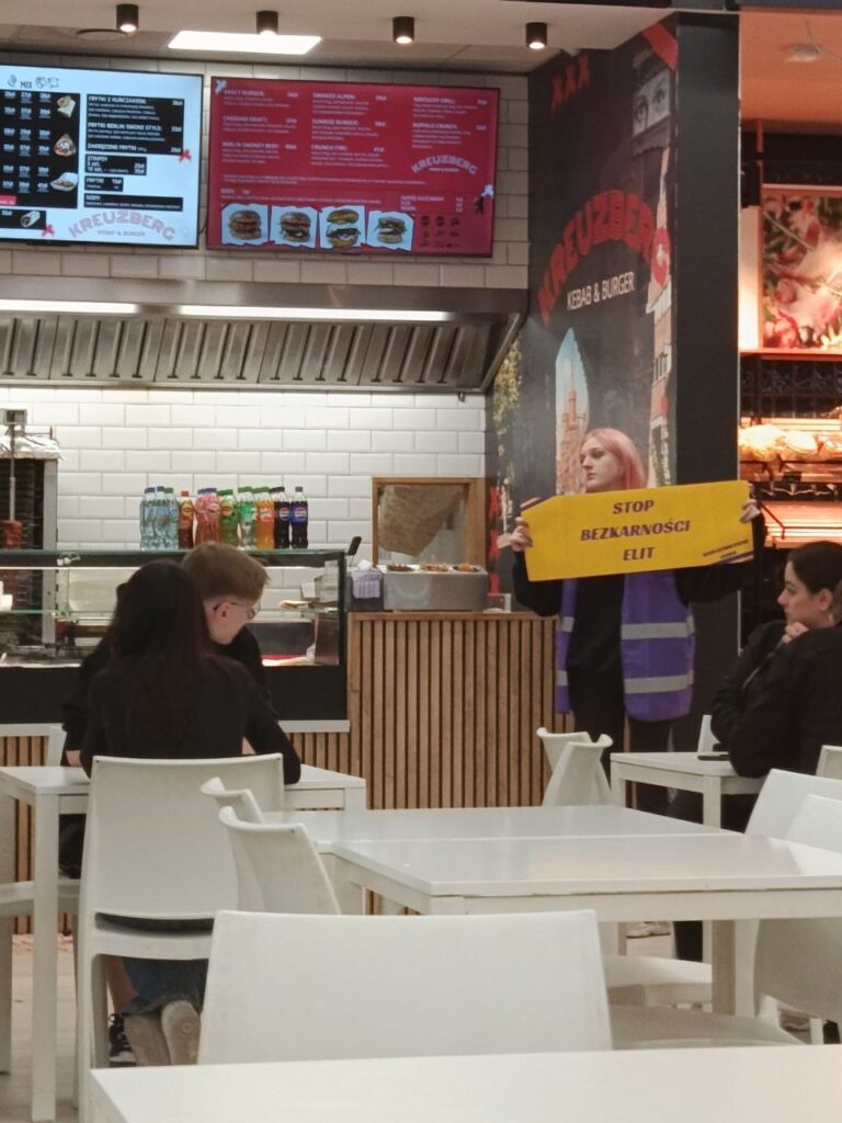 Kobieta z różowymi włosami trzyma transparent z napisem „STOP BEZKARNOŚCI ELIT” w lokalu gastronomicznym.