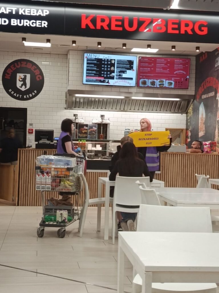 Kobieta z różowymi włosami trzyma transparent „STOP BEZKARNOŚCI ELIT” w lokalu gastronomicznym Kreuzberg, obok stoiska z jedzeniem.