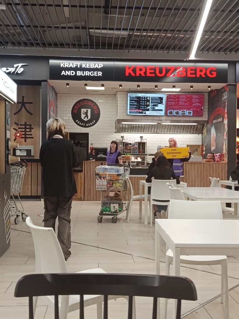 Kobieta w fioletowym fartuchu obsługująca klientów w strefie gastronomicznej z szyldem "KRAFT KEBAP AND BURGER".