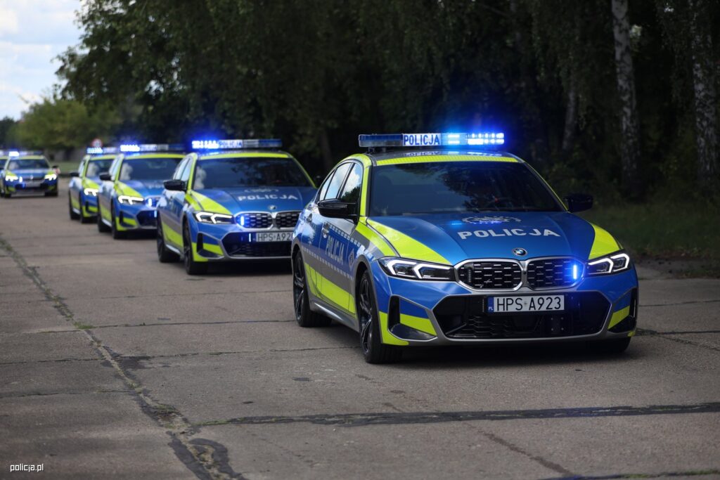 Grupa policyjnych samochodów marki BMW z włączonymi sygnalizatorami świetlnymi, ustawionych na asfaltowej drodze.