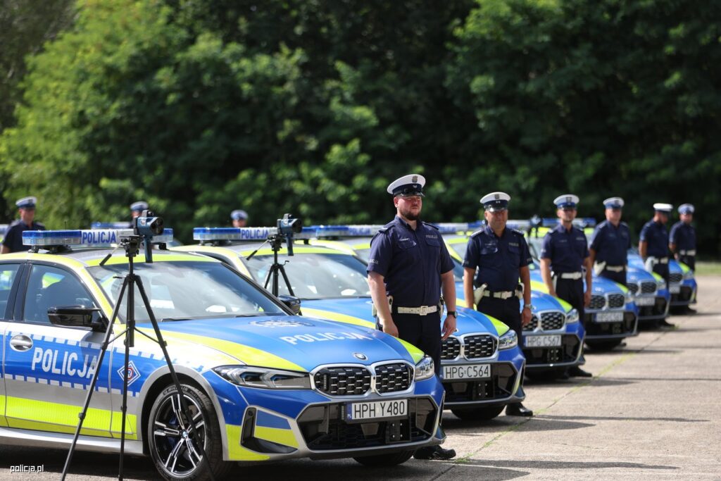 Grupa policjantów w mundurach stoi w szeregu obok nowych radiowozów z niebieskimi sygnalizatorami.