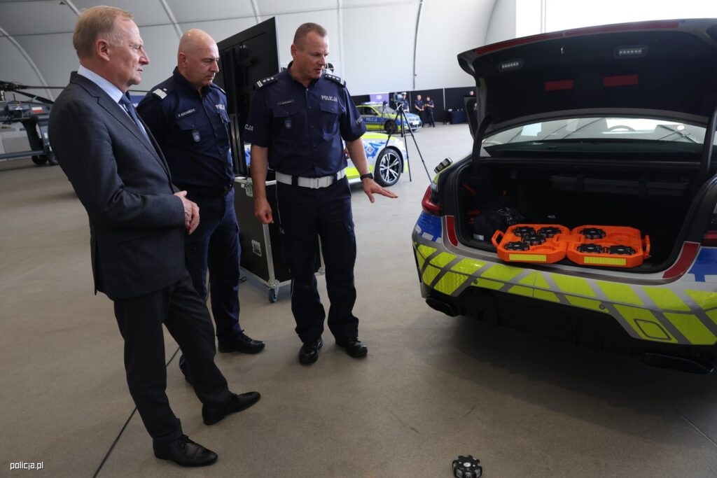 Mężczyzna w garniturze i dwóch policjantów w mundurach omawiają sprzęt w bagażniku radiowozu.