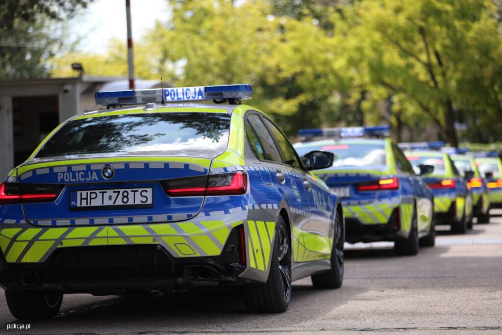 Policyjne samochody osobowe w niebiesko-żółtej kolorystyce ustawione w rzędzie na ulicy, w tle zieleń drzew.