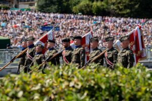 Osiemnastu żołnierzy w mundurach wojskowych, niosących sztandary, maszeruje przed tłumem widzów na defiladzie.