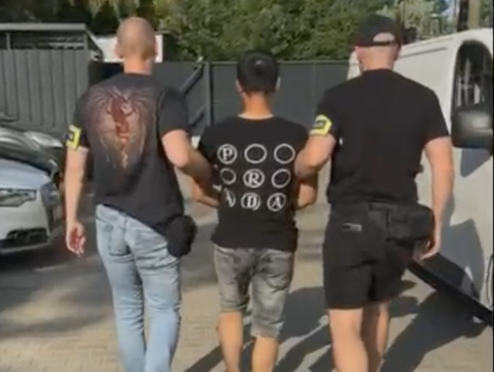 Mężczyzna w t-shircie z okręgami prowadzony przez dwóch funkcjonariuszy w czarnych koszulach w otoczeniu samochodów.