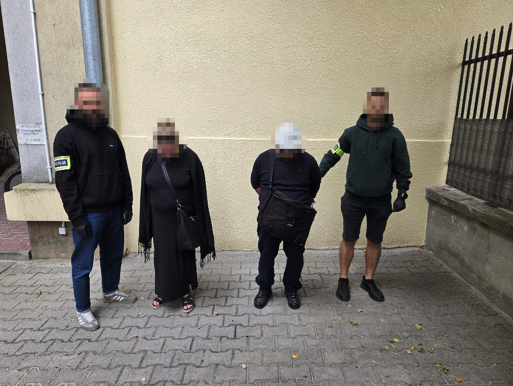 Czwórka osób stoi na chodniku, jedna z nich trzyma mężczyznę w czarnym ubraniu, wszyscy mają na sobie ciemne odzież.