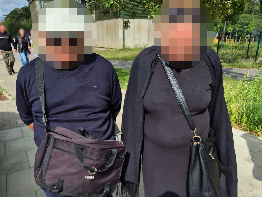 Dwoje ludzi stoi na chodniku, mężczyzna w granatowej bluzie z torbą na ramieniu i kobieta w czarnej sukience z torebką.
