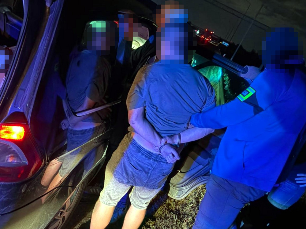 Mężczyzna w kajdankach jest zatrzymywany przez policjanta przy otwartym samochodzie w nocy.