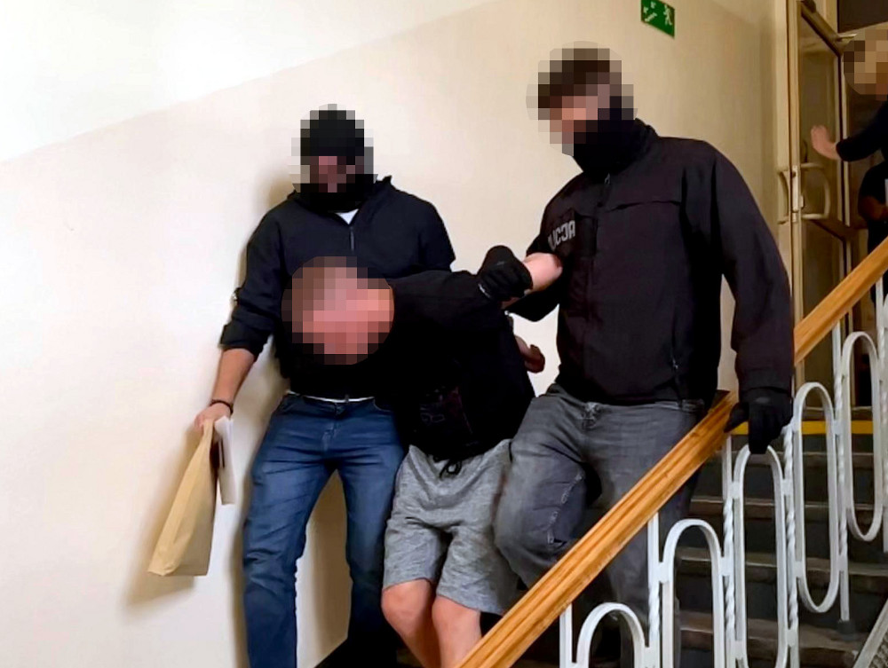 Trzech funkcjonariuszy w maskach eskortuje mężczyznę w krótkich spodniach schodami w budynku.