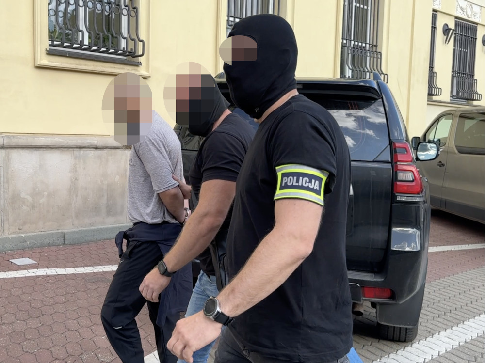 Trzech mężczyzn w maskach jest prowadzonych przez funkcjonariuszy policji na ulicy, obok czarnego SUV-a.