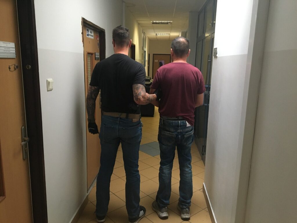 Mężczyzna w czarnej koszulce prowadzony przez policjanta w korytarzu budynku, obok drzwi do biura.