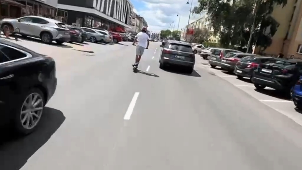 Mężczyzna w białej koszulce jedzie na hulajnodze po ulicy, obok zaparkowanych samochodów.