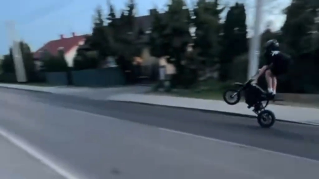 Młody mężczyzna wykonuje wheelie na małym motocyklu na ulicy, obok domów i drzew.
