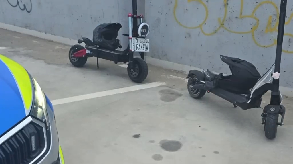 Dwa elektryczne hulajnogi zaparkowane obok siebie na parkingu, w tle widoczna szara ściana z graffiti.