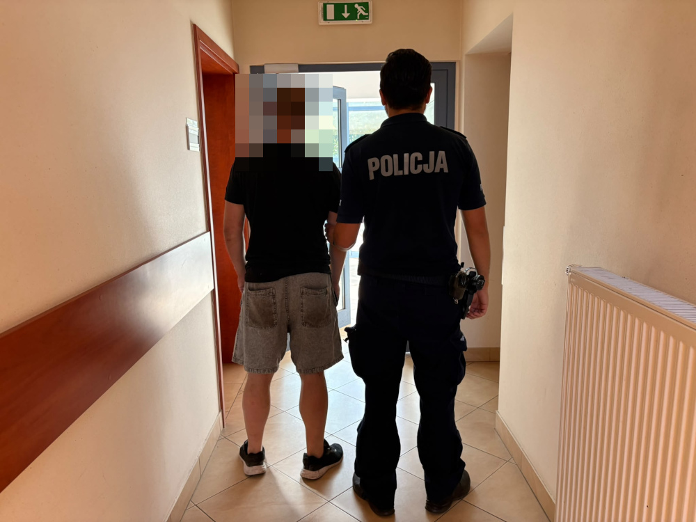 Mężczyzna w krótkich spodniach prowadzony przez policjanta korytarzem w budynku.