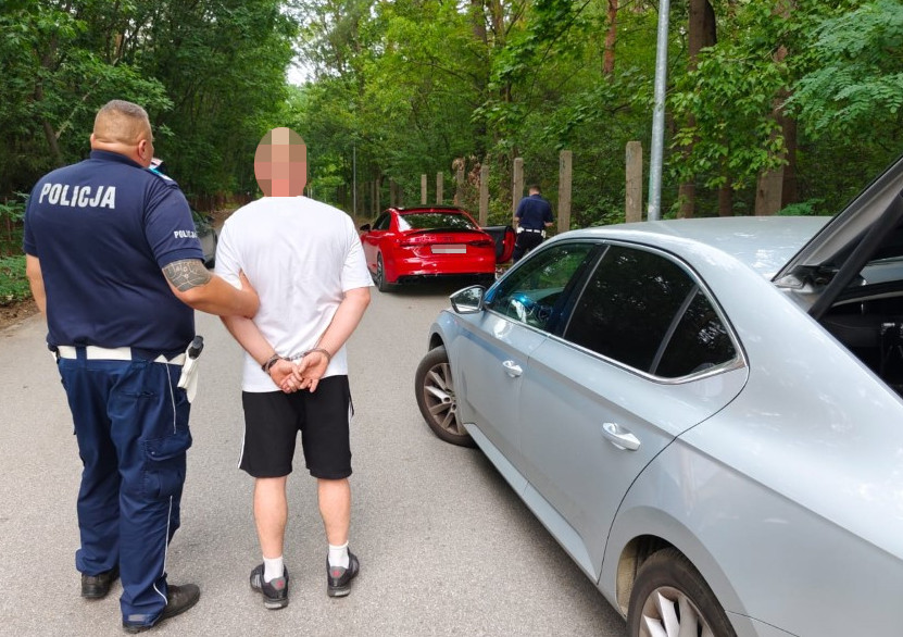 Policjant zatrzymuje mężczyznę w białej koszulce na leśnej drodze, obok stoi czerwony samochód sportowy.