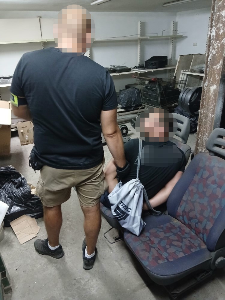 Funkcjonariusz policji trzyma mężczyznę w ciemnym ubraniu siedzącego na fotelu w pomieszczeniu z porozrzucanymi przedmiotami.