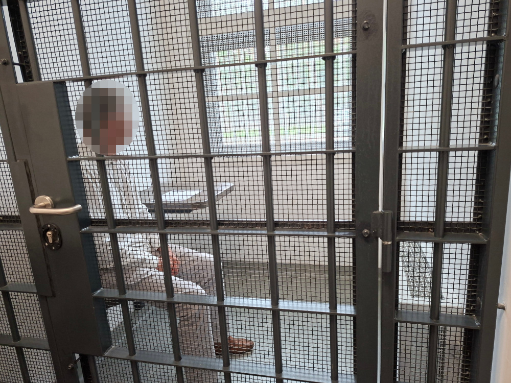 Mężczyzna w białym ubraniu siedzący w celi, z widokiem na zewnętrzny świat przez metalową kratę.
