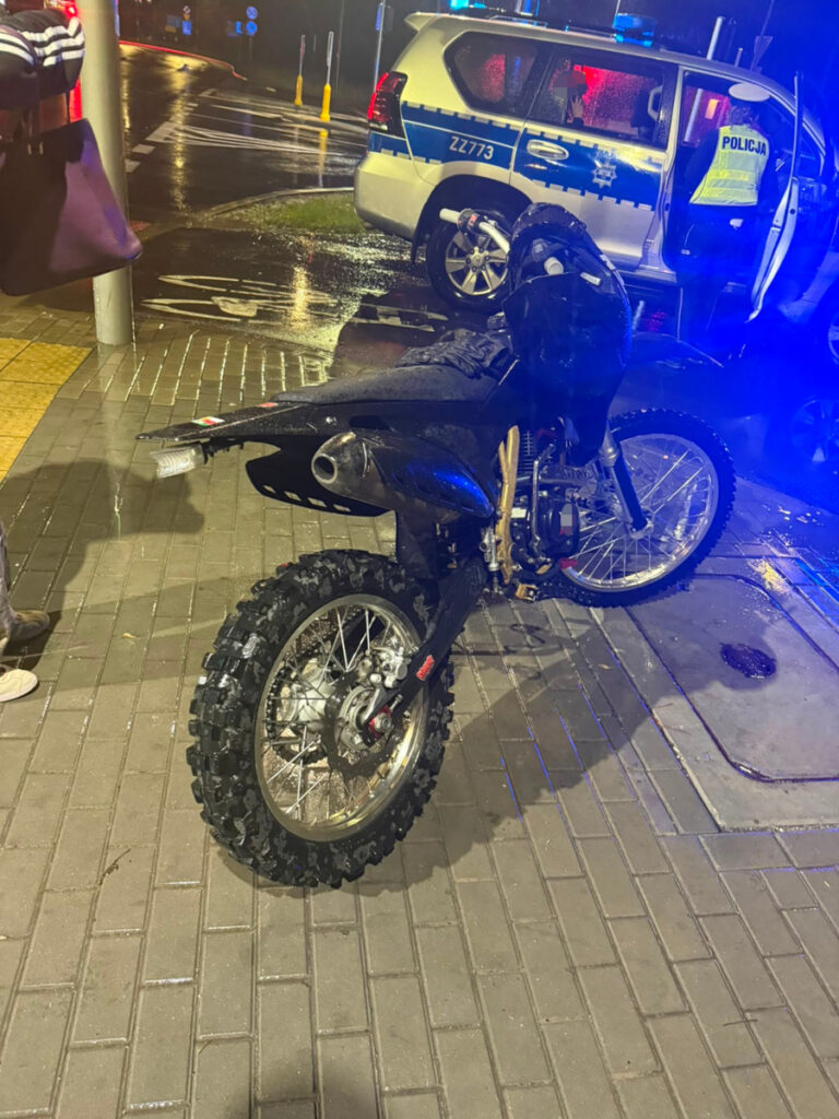 Motocykl terenowy zaparkowany na mokrej nawierzchni, obok radiowozu policyjnego z włączonymi sygnałami świetlnymi.