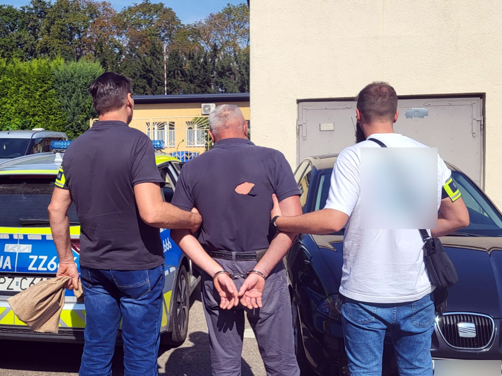 Mężczyzna z rękami związanymi z tyłu jest prowadzony przez dwóch strażników miejskich na parkingu.