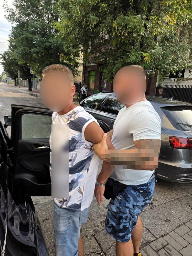 Mężczyzna w koszulce z motywem roślinnym jest zatrzymywany przez funkcjonariusza w białym t-shircie na ulicy.