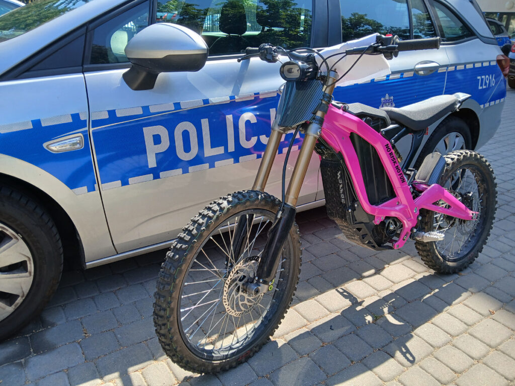 Elektryczny motocykl w różowym kolorze stoi obok policyjnego samochodu z widocznym napisem "POLICJA".