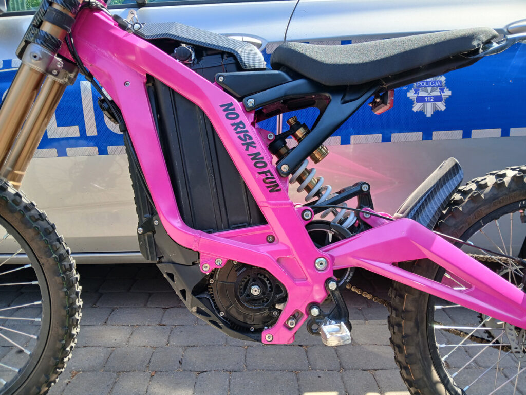 Różowy elektryczny motocykl o sportowym designie stoi na chodniku obok radiowozu policji. Na ramie widnieje napis "NO RISK NO FUN".