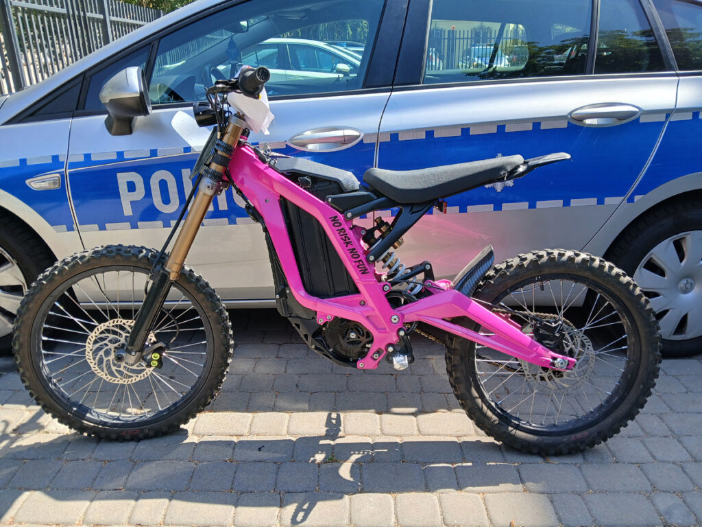 Elektryczny motocykl w różowym kolorze stoi na chodniku obok radiowozu policyjnego.