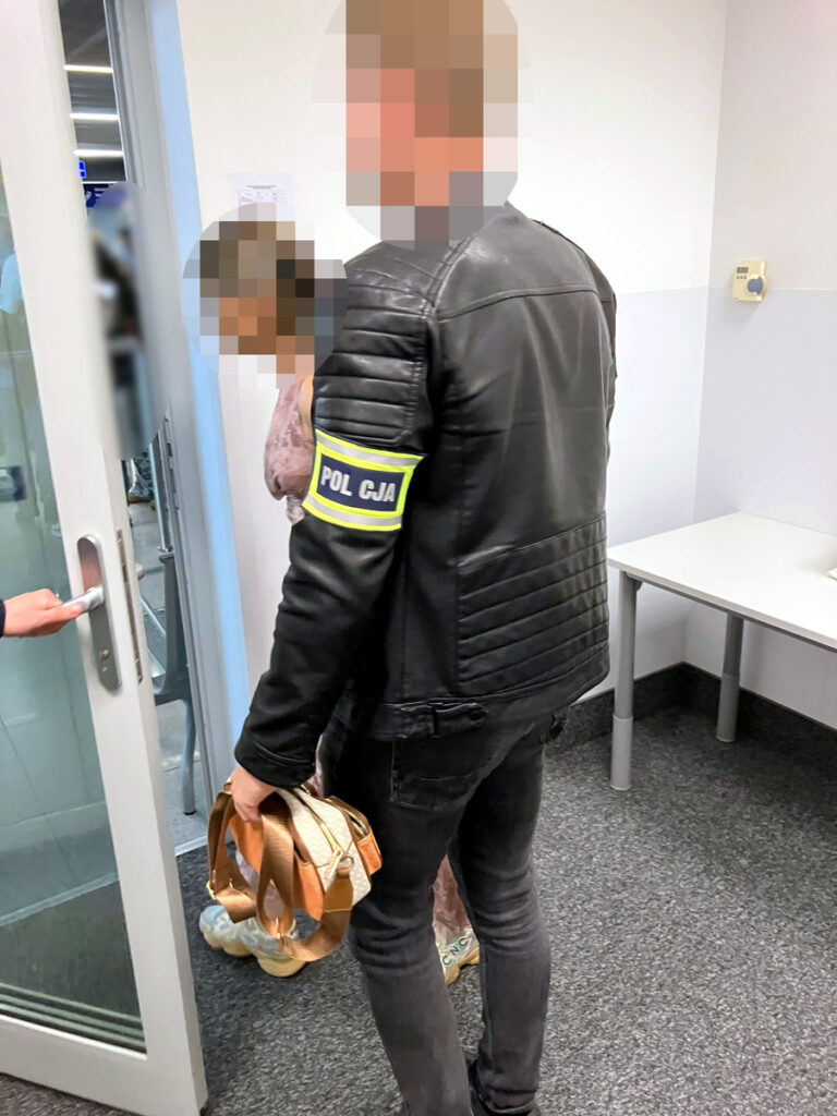 Funkcjonariusz policji w skórzanej kurtce prowadzi osobę z torbą w kierunku drzwi w nowoczesnym pomieszczeniu biurowym.