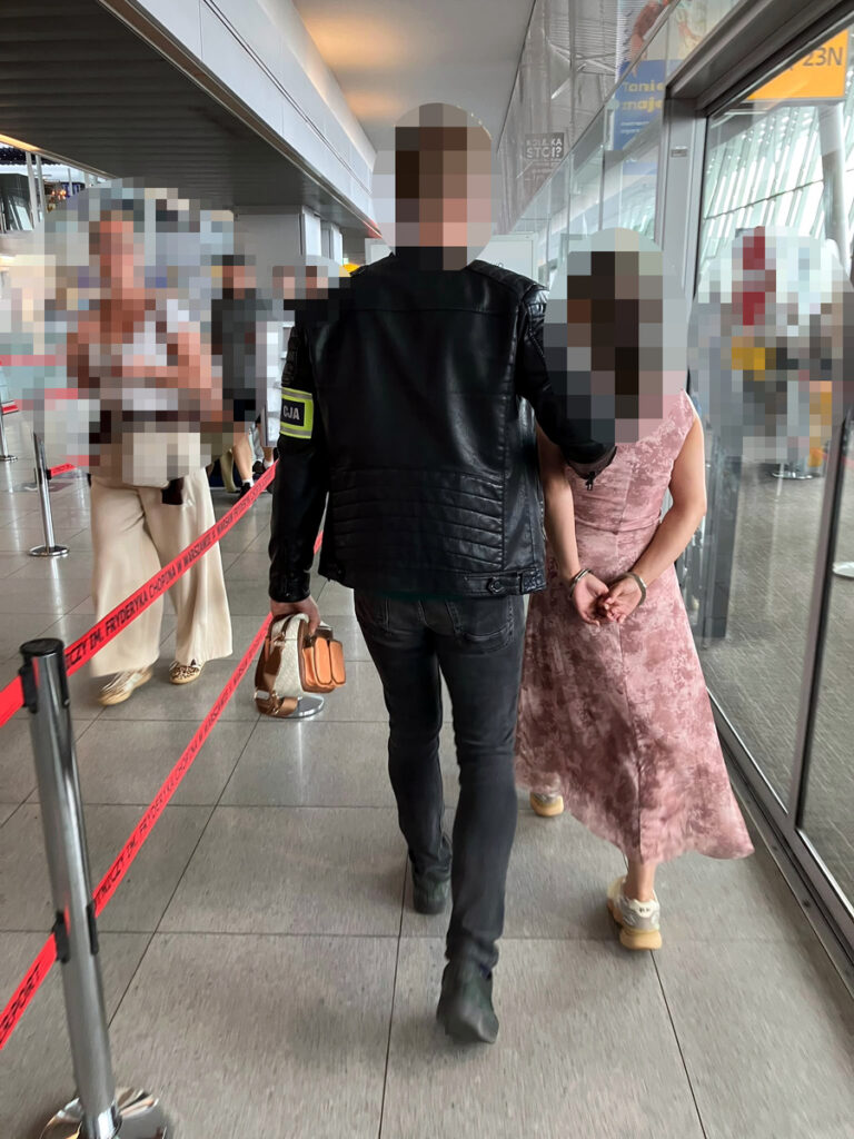 Mężczyzna w skórzanej kurtce prowadzi kobietę w różowej sukience, trzymając ją za rękę w przestrzeni lotniska.